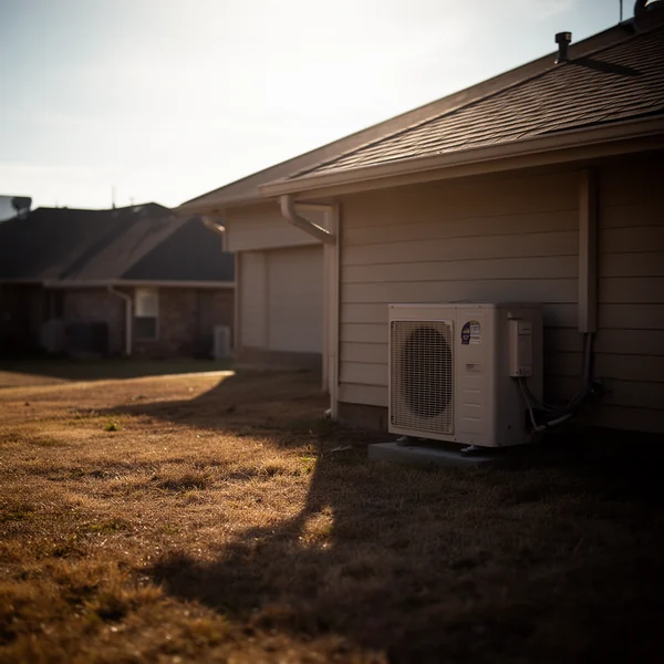 Heat shimmer over a suburban rooftop in late afternoon, a condenser unit at the side of the house, dry grass and hard shadows