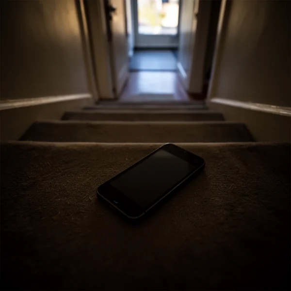 A phone lying on a stair tread in a dim hallway, screen dark, cool morning light from a doorway above